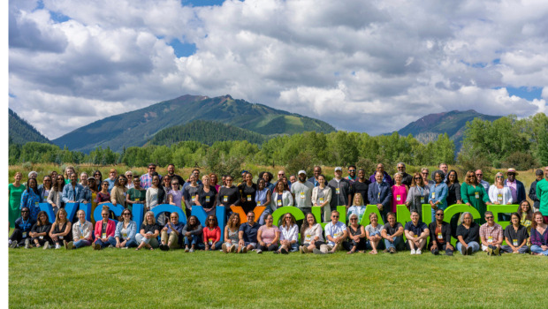 A large group of event participants stand and sit together, smiling. They are on a grassy area and behind them are the rocky mountains and a cloudy sky.